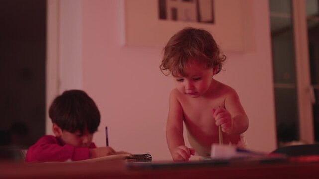Toddler reaching across table and scribbling in notebook next to older brother studying, lively family scene blending play and homework at home