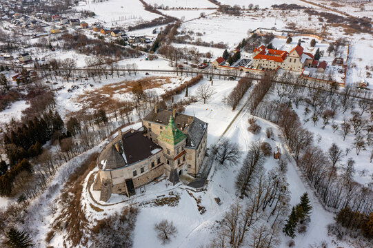 Aerial view of Olesko Castle perched atop a snow-dusted hill, its ancient walls contrasting with the white landscape and the distant monastery, Olesko, Lviv Oblast, Ukraine.