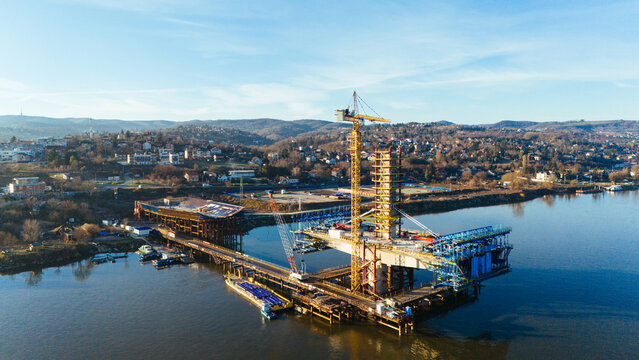 Aerial view of a construction site with a crane and bridge supports rising from the water, reflecting the crisp morning light, Novi Sad, Vojvodina, Serbia.