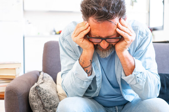 Sad and frustrated 50s man suffering from stress, depression at home, sitting on sofa, touching head, feeling headache, migraine, dealing with loss, grief, trauma. Nervous breakdown, crisis concept