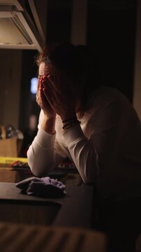 Woman standing at kitchen counter holding head in hands, tired expression, late evening stress and domestic exhaustion moment