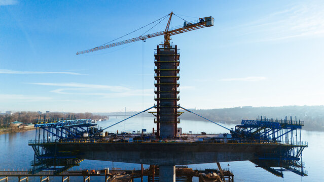 Aerial view of a colossal bridge pier rises from the water, crowned by a yellow crane against the vast sky, Novi Sad, Vojvodina, Serbia.