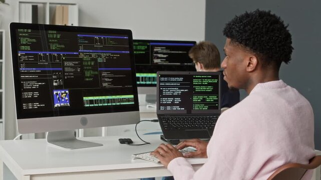 Side view of young adult Black male software engineer coding on desktop computer troubleshooting bugs in scripts at workstation in modern open plan office
