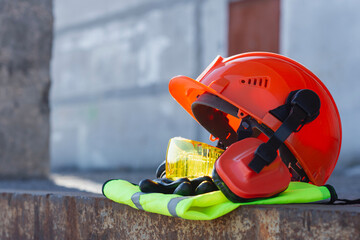 Orange hard hat with protective goggles, earmuffs and high visibility vest against industrial background. Personal protective equipment for construction workers and industrial safety.