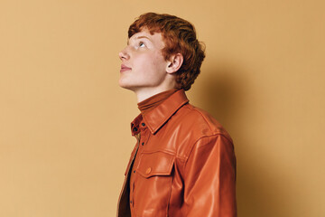 Young man with red hair wearing brown leather jacket looking upward on beige background. Profile portrait with thoughtful expression and trendy casual style in studio light. © SHOTPRIME STUDIO