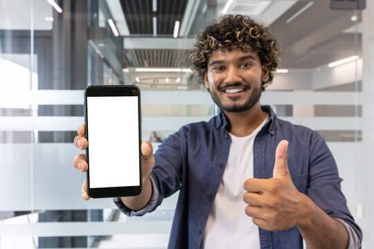 Portrait of a young smiling Indian man in the office, holding and showing the camera a white phone screen, showing a super sign with his finger