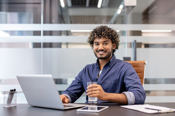 Fototapeta premium Portrait of a young Indian man sitting at a desk with a laptop, holding a glass of water in his hand and looking at the camera with a smile