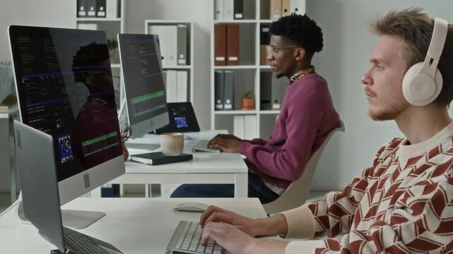 Rack focus of young adult male developer with headphones on programming on desktop computer next to desk of focused colleague in open plan office