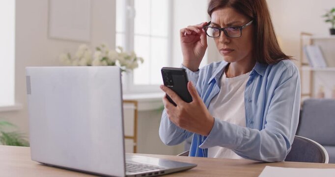 Woman with poor eyesight trying to read text on smartphone screen, taking off and putting on glasses to focus. Presbyopia, myopia and eyestrain of lady with eyeglasses sitting at laptop at home desk