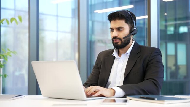 Man corporate operator in headset working in customer support service on helpline telesale talking using laptop in business office. Agent of call center in suit advises client by answering questions.