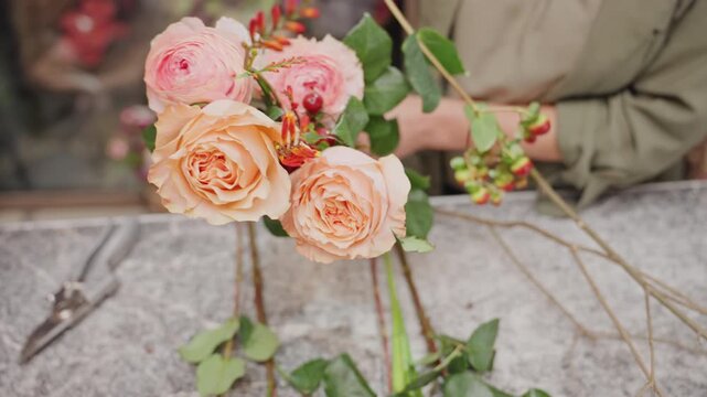 Florist assembling bridal bouquet on workbench, scissors and stems scattered, steady hands tying stems with ribbon, hypericum berries adding texture, natural light highlighting process