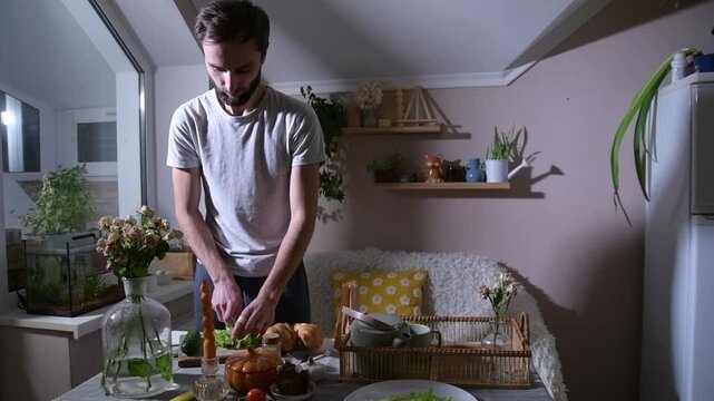 Man preparing croissants for breakfast in bed, filling them with fresh lettuce leaves at the kitchen table. Cozy morning lifestyle scene showing a thoughtful homemade meal and caring gesture.
