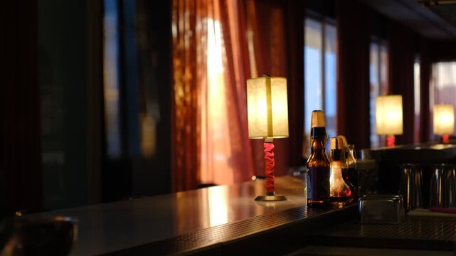 Atmospheric dimly lit bar counter with vintage lamps, beer bottles, and red curtains at sunset.