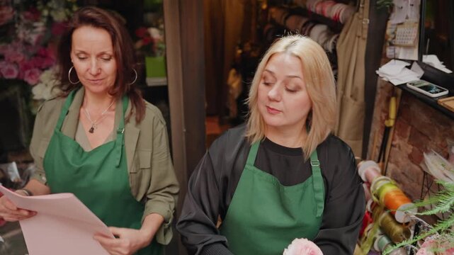 Two caucasian florists wrapping pink bouquet together at counter. Cooperative shop scene with mentor and apprentice exchanging paper and ribbon, green aprons, scissors and twine, bouquet of roses