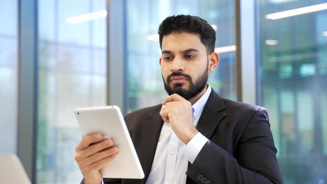 Thoughtful businessman in formal suit using browsing digital tablet sitting in business office. Serious manager thinks about task, works in app, typing message or chatting online with client. Close up