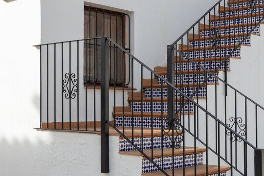 Traditional Spanish staircase featuring terracotta treads and blue-and-white patterned ceramic tiles. The whitewashed exterior is accented by wrought-iron railings and matching window security grilles