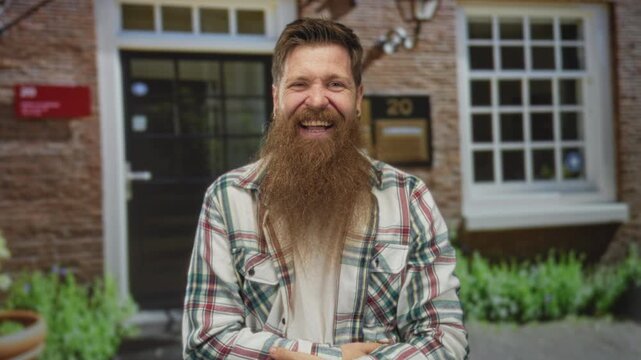 Man in plaid shirt with long beard covering face with hand at building entrance in daylight; frustration.