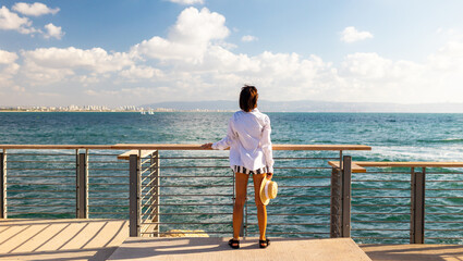Slender woman with a straw hat in her hands looks at the ocean. Summer vacation by the sea. Freedom and relaxation on the seashore.