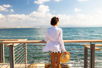 Slender woman stands on the embankment and looks at the ocean. Summer vacation by the sea. Freedom and relaxation on the seashore.