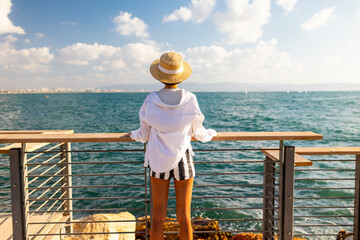 Slender woman stands on the embankment and looks at the ocean. Summer vacation by the sea. Freedom and relaxation on the seashore.