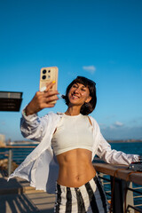 Female blogger shoots content for social networks and waves at the camera. Smiling tourist takes a selfie on a mobile phone against the backdrop of the sea.