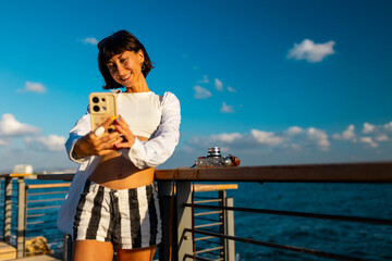 Attractive smiling tourist taking a selfie on her mobile phone against the backdrop of the Mediterranean Sea. The girl enjoys her vacation and shoots content for social media.