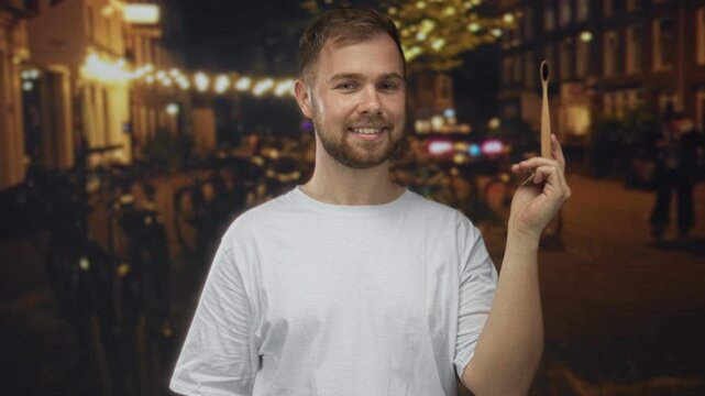 Smiling man holding bamboo toothbrush in hand on city street at night under string lights; sustainable oral care happiness.