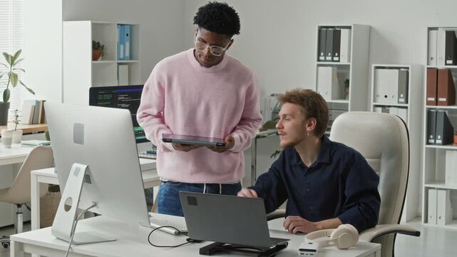 Young adult Caucasian male senior developer pointing at computer screen guiding junior Black male colleague with tablet through code debugging while collaborating at workdesk in IT office