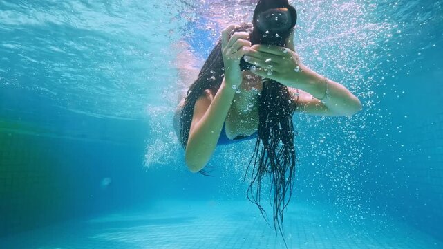 Young photographer wearing sunglasses and a blue swimsuit taking photos underwater in a swimming pool while many bubbles are coming out of her nose