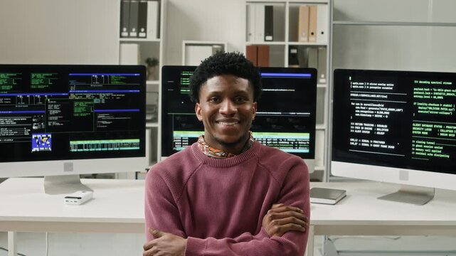 Medium portrait shot of smiling young African American male cybersecurity engineer sitting at workstation with multiple computer screens then turning to camera with arms crossed in modern IT office