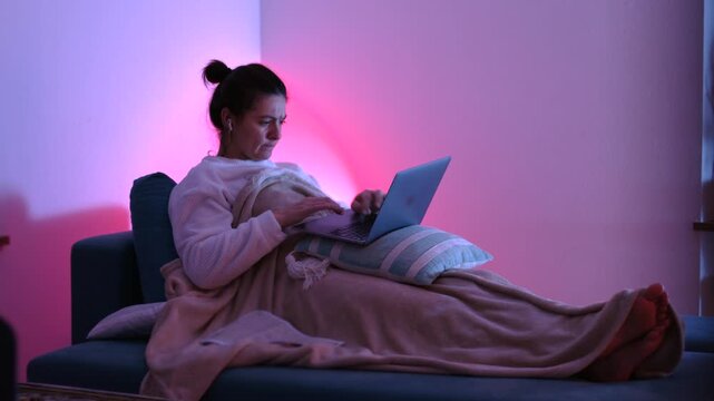 Woman using laptop while reclining on couch with blanket at night, managing remote workload and staying connected through digital platforms from home
