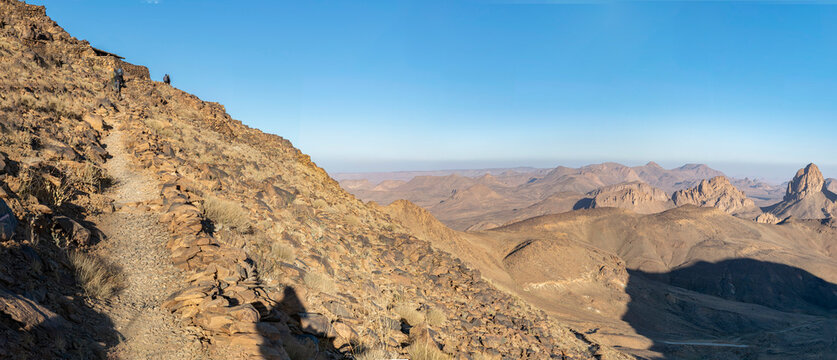 Blurred people climbing the Assekrem elevated path over the granite and volcanic Hoggar Mountains in the Atakor near Tamanrasset, jagged peaks casting shadows in a sunny day under a clear blue sky.
