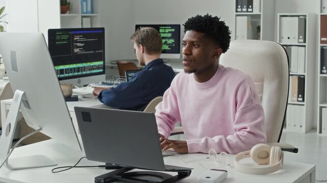 Medium shot of focused young adult Black male full stack developer typing on keyboard at workstation with laptop and desktop computer while debugging errors in programs in open plan office