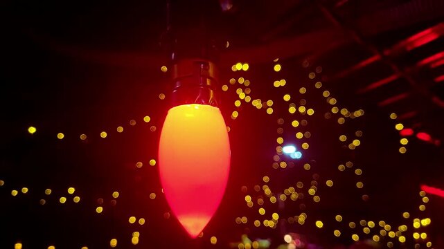 Slow zoom out on a red C7 Christmas light bulb illuminated against a dark background with blurred yellow festive lights.