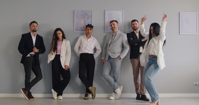 Energetic dance of excited woman dancing in front of team of sad employees standing together by office wall. Young woman performing happy expressions of winner, exotic dance to music to celebrate win
