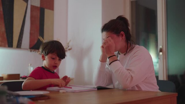 Mother supervising son while he writes in notebook at dining table, candid home education scene capturing attention, guidance and shared responsibility