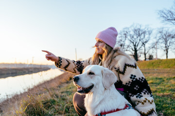 Focused female, blonde Caucasian, pointing direction while sitting with golden retriever outdoors © Iryna