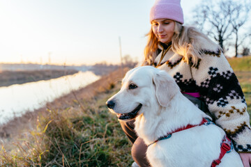Cheerful female, blonde Caucasian, crouching with golden retriever on riverbank at sunrise © Iryna