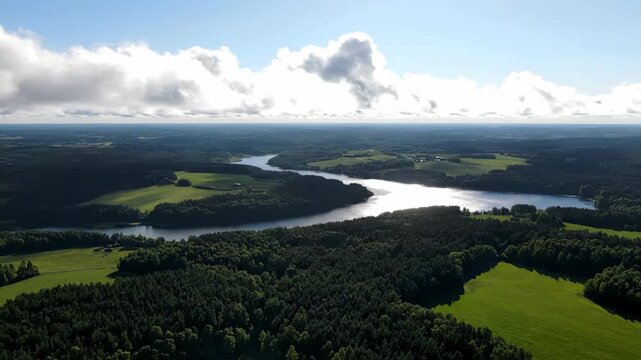 Aerial view of a serene landscape, with a winding river and expansive forest under a cloudy sky. Patches of sunlight illuminate verdant fields