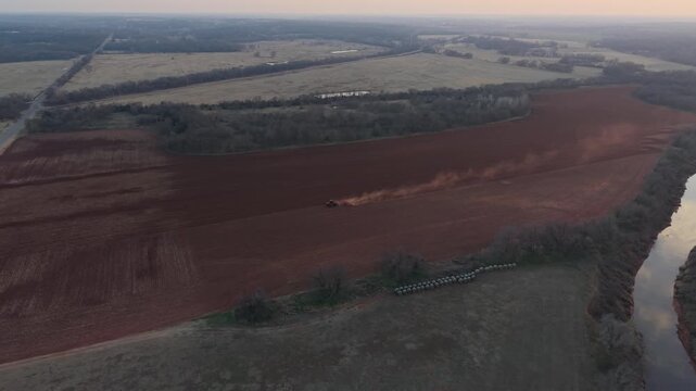 Drone aerial pulling back while tracking a tractor plowing red dirt farmland beside a winding creek in rural Oklahoma agricultural landscape.