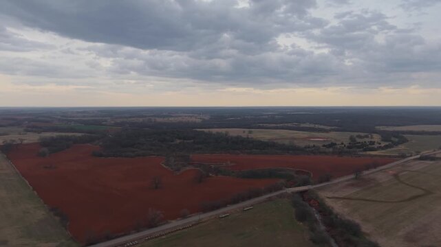 Drone aerial pulling back and orbiting over red dirt farmland, rural road bridge, and winding creek beneath dramatic storm clouds in Oklahoma countryside.