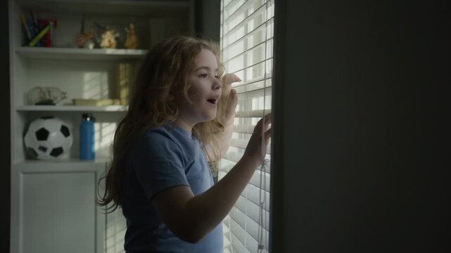 Child looking out the window with an expression of surprise. Kid with excitement near window. Child showing a moment of wonder by the window. Amazed kid looking through the blinds in soft light.