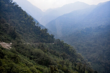 Fototapeta premium Winding mountain road through dense Himalayan forest with misty blue hills and steep valley landscape in North Sikkim.
