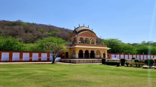 A yellow historic pavilion with elegant arches stands in a landscaped garden in Jaipur, backed by green hills. The ornate structure reflects traditional Rajput architecture under a clear blue sky.