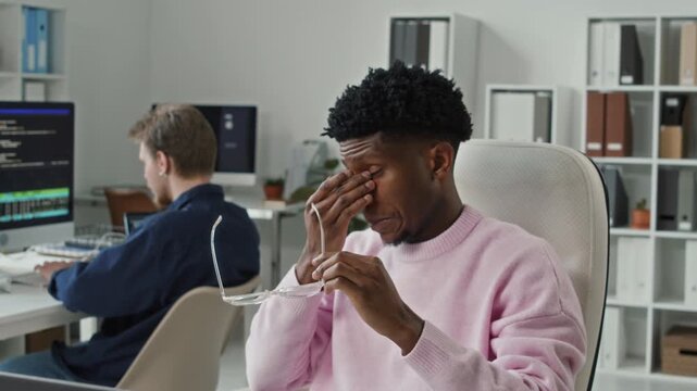 Dolly away shot tired young adult Black male programmer taking off glasses to rub strained eyes then unwinding with arm stretch while working on computer in open space office with colleagues