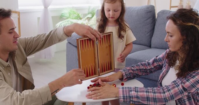 Family home daughter and parents play board game. Mother and father sit together at table, guiding child girl through wooden backgammon set and childhood turn-taking. Happy family time and leisure.