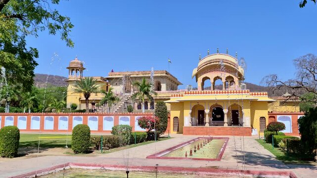 A beautiful heritage garden in Jaipur with fountains leading toward a yellow pavilion with ornate arches. Palm trees, a watchtower, and surrounding hills highlight the grandeur of Rajput architecture.