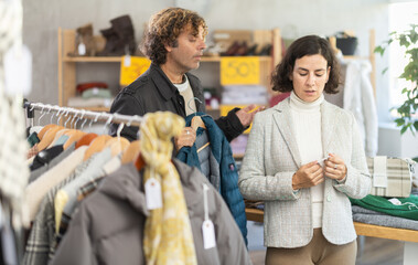 While shopping, woman buyer looks at products in store and selects jacket or coat, irritated man is...