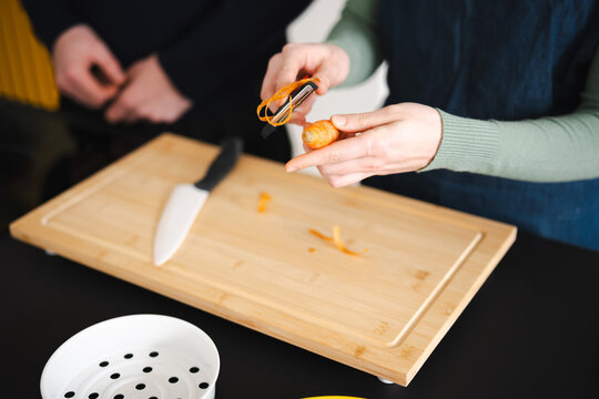 Two people prepare food in a kitchen, one person peels a carrot on a wooden cutting board with a knife nearby.