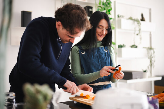 A couple prepares vegetables together in a bright kitchen. The man slices carrots on a cutting board while the woman peels another carrot.